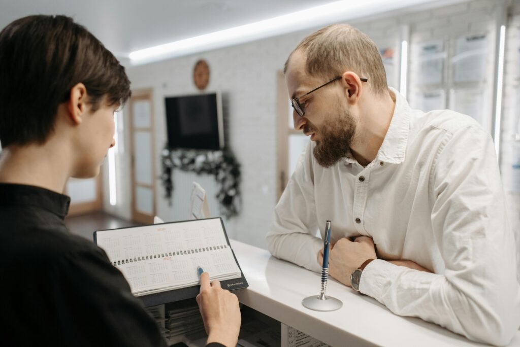 A receptionist and client converse over an appointment book at a clinic reception desk.